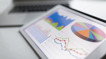 Ansicht eines Tablets mit grafischen Darstellung verschiedener Statistiken. Im Hintergrund ist verschwommen ein Laptop zu sehen.