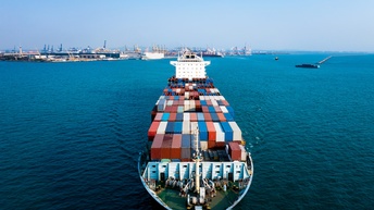 Ein Frachtschiff mit vielen Containern, das auf einem Gewässer fährt. Im Hintergrund ist ein Hafen mit vielen Hafenkränen