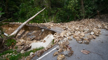Zerstörter Straßenabschnitt in Kurve verlaufend voller Steingeröll, ein entwurzelter umgekippter Baum liegt über der Straße, im Hintergrund grüne Sträucher und Bäume