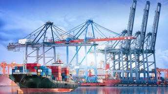 Im Vordergrund ist ein Transportschiff mit vielen Containern im Wasser. Dahinter ist eine Hafenanlage mit weiteren Schiffen, Containern und anderen Utensilien für die Logistik
