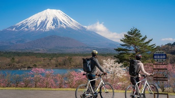 Zwei Personen mit Fahrrad, Schutzhelm und Rucksack stehen auf einer Straße und blicken in Richtung des Sees Shojiko am Fuße des dahinterliegenden Mount Fuji 