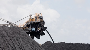 An einem abgeflachten, dunklen Hügel aus sehr dunklem Material ist am Rand ein großer Schaufelradbagger, der das Material aushebt und zur Seite schaufelt.