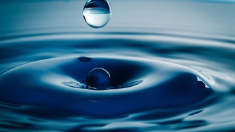 Nahaufnahme einer Wasseroberfläche, in die ein Wassertropfen fällt, wodurch die Oberfläche gebrochen wird und Wellen schlägt.