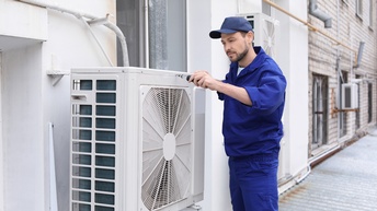 Person in blauer Arbeitskleidung mit Kappe steht auf Dachvorsprung zur Hauswand neben Fenster und schraubt an Außengerät einer Klimaanlage