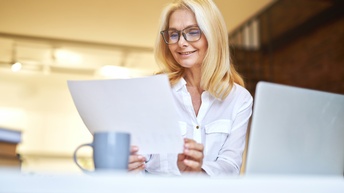 Lächelnde Person mit Brillen an Schreibtisch vor aufgeklappten Laptop sitzend hält Dokument in Händen und blickt darauf