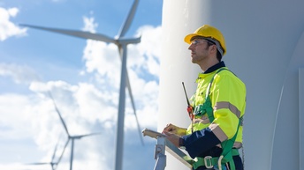 Seitliche Aufnahme einer Person mit Helm, Schutzbrille und Sicherheitsjacke, die ein Tablet hält. Ihr Blick geht leicht nach oben. Hinter der Person stehen mehrere, versetzte Windräder unter blauem, wolkenverhangenem Himmel.