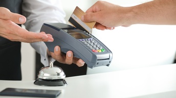 Person hält Kreditkarte zu einem kontaktlosen Bezahlgerät, daneben steht eine Klingel einer Rezeption