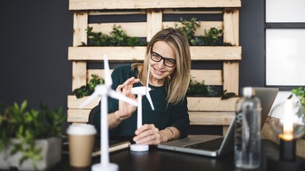 Person mit Brillen sitzt lächelnd an Schreibtisch und blickt auf Modelle von Windrädern, vor ihr aufgeklappter Laptop