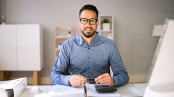 Lächelnde Person mit Brille sitzt an einem Schreibtisch und arbeitet mit aufgeschlagener Mappe, Taschenrechner, im Hintergrund befinden sich Regale und Ablagen