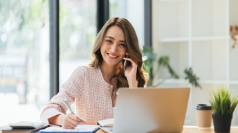 Person mit langen Haaren sitzt bei einem Schreibtisch telefoniert mit einem Smartphone, während mit einem Stift Notizen gemacht werden und arbeitet mit einem Laptop in einem modernen, hellen Büro