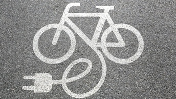Detailansicht Asphaltstraße mit weißem aufgemalten Fahrradzeichen, Linie des Fahrrads verläuft in Zeichen für Kabel mit Stromstecker
