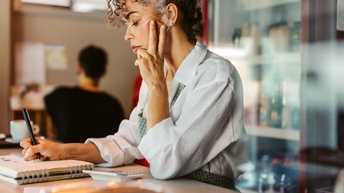 Seitliche Aufnahme einer Person mit einer Schürze, die an einem erhöhten Tresen steht und vor sich einen Notizblock hat und einen Stift hält. Daneben steht ein Taschenrechner