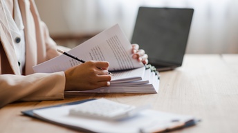 Seitliche Aufnahme einer Person, die sich durch einen Papierstapel arbeitet und einen Bleistift hält. Neben der Person sind außerdem ein Klemmbrett, ein Taschenrechner und ein aufgeklappter Laptop