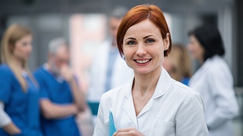 Porträt einer Person aus dem medizinischen Personal einer Einrichtung. Die Person trägt ein weißes Oberteil und lächelt zufrieden. In der Hand hält sie eine blaue Mappe. Im Hintergrund ist weiteres medizinisches Personal zu sehen.