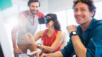 Drei lächelnde Personen an Schreibtisch stehend und sitzend, eine Person trägt VR-Brille und greift mit Händen in die Luft