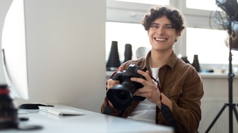 Porträt einer lächelnden Person auf Stuhl an Tisch sitzend hält Spiegelreflexkamera in Händen, im Hintergrund verschwommen Scheinwerfen und Objektive