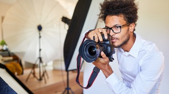 Eine Person mit Brille, in einem weißen Hemd beugt sich mit dem Oberkörper und dem Kopf leicht nach vorne. Sie hält mit beiden Händen seitlich eine Kamera. Die Person blickt geradeaus