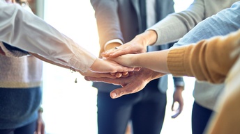 Detailaufnahme der Hände von fünf Personen, die in der Mitte aufeinander gelegt werden. Im Hintergrund ist ein Fenster, das Licht auf die Hände wirft, sodass diese durch den Lichteinfall stark belichtet sind. 