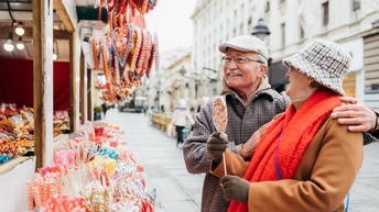Zwei lächelnde Personen mit Brillen und Kopfbedeckungen stehen an Markthütte voller Süßwaren, eine Person hält großen Lutscher in der Hand