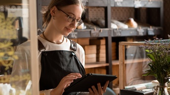 Person mit Brille in Arbeitskleidung blickt auf ein Tablet hinter einer Verkaufstheke