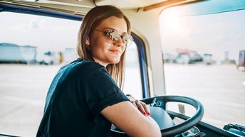 Portrait einer sanft lächelnden Person mit Sonnenbrillen in Fahrerkabine eines Lastkraftwagens sitzend, die Arme verschränkt am Lenkrad abstützend, im Hintergrund verschwommen weitere parkende Lastkraftwagen