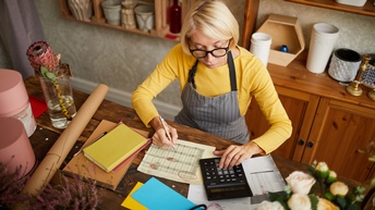 Person mit kurzen blonden Haaren, Brille, gelben Pullover und Arbeitsschürze sitzt an einem Tisch und blickt auf einen Taschenrechner während ein Stift auf einem Dokument gehalten wird, daneben stehen Blumen, Vasen und Notizbücher, Blick von oben