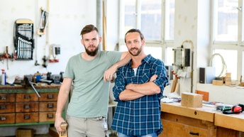 Zwei Personen mit Bart stehen zusammen und blicken in die Kamera, im Hintergrund zeigt sich eine Werkstätte