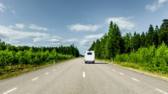 Rückenansicht eines Wohnmobils, das auf einer zweispurigen Straße fährt. Links und rechts von der Straße sind eine grüne Wiese und grüne Bäume