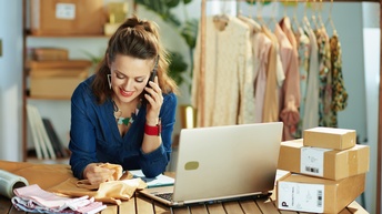 Lächelnde Person mit Modeschmuck neigt sich über Tisch und tastet mit einer Hand Stoff, hält mit anderer Hand Smartphone ans Ohr, daneben am Tisch weitere Stoffe, aufgeklapptes Notebook, Kartonagenstapel, im Hintergrund  behängte Kleiderstangen und Regal