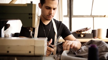Person sitzt an Tisch und schneidet mit Schere Stoff, im Vordergrund verschwommen im Ausschnitt Nähmaschine, im Hintergrund verschwommen emailliertes Fenster und Tisch mit Zwirnen und Stoffmustern