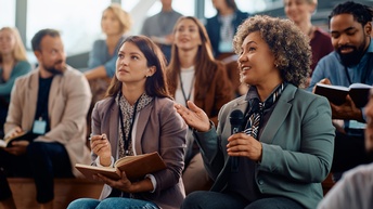 Mehrere Personen in Bürokleidung sitzen in einem Auditorium und blicken nach vorne. Die Person rechts unten im Bild hält in der linken Hand ein Mikrofon