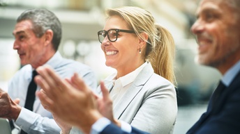 Drei Personen in Businesskleidung sitzen nebeneinander und applaudieren.