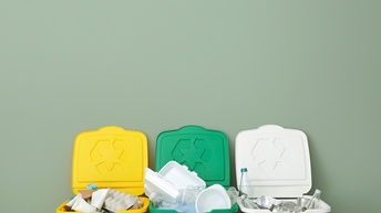 Vor einer grünlichen Wand stehen nebeneinander drei geöffnete Mülltonnen. In jeder Mülltonne ist unterschiedlicher Müll. Ganz links Karton, in der Mitte Plastik und rechts Glas und Metall