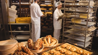 In einer Bäckerei: im Vordergrund Körbe und Backblech mit Backwaren, im Hintergrund schieben zwei Personen Bleche in Regale 
