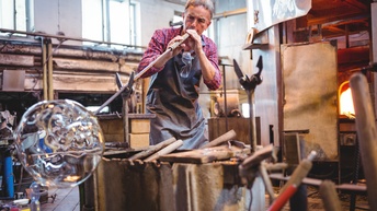 Person in Schürze in Werkstatt mit Brennofen stehend bläst durch Rohr Glaskugel, ringsum verstreut Werkzeuge