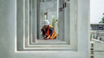 Person in oranger Warnweste mit weißem Schutzhelm vermisst Betonteil, das in selber Ausfertigung mehrfach davor in Reihe steht