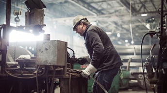 Person mit Schutzhelm und Schutzhandschuhen steht in großer Werkhalle an  Maschine und hält Metallblatt unter Gerät