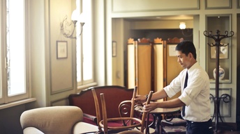 Person mit dunklen Haaren, Hemd und Krawatte ordnet Stühle in einer Hotel Lounge