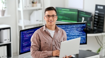 Lächelnde Person mit Brille hält aufgeklapptes Notebook in Händen, im Hintergrund verschwommen Computerbildschirme auf denen Quellcodes aufscheinen 