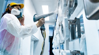 Person mit weißem Schutzanzug und Atemmaske sprüht Farbe auf ein Objekt in einer Werkstätte