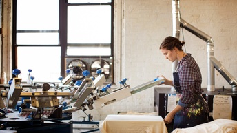 Person in Schürze mit Farbflecken bedient Siebdruckmaschine, im Hintergrund Metallrohre und Fenster
