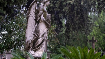 Steinskulptur eines Engels in Seitenansicht zwischen Oleanderpflanze und Palmen, im Hintergrund Nadelbäume und Steinkreuz