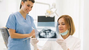 Zwei Personen in Zahnarztpraxis: eine stehende Person in blauem Arbeitsgewand macht Notizen und blickt auf Röntgenbild eines Gebisses, das andere Person in weißem Arbeitsgewand mit Handschuhen hoch hält