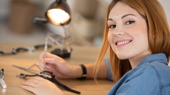 Lächelnde Person im Fokus, im Hintergrund verschwommen Tisch, an dem Person Uhr mit Werkzeugen unter Lampe bearbeitet