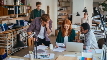Fünf Personen in einem Büro. Drei Personen beschäftigen sich gemeinsam mit Unterlagen und einem Laptop und sitzen bzw. stehen an einem Tisch. Im Hintergrund befinden sich zwei weitere Personen