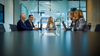 Sechs Personen in Bürokleidung sitzen um einen großen Tisch. Vor zwei Personen steht ein aufgeklappter Laptop. Die Personen blicken sich gegenseitig an. Hinter ihnen ist eine Glaswand, auf der die Kontinente der Erde sind