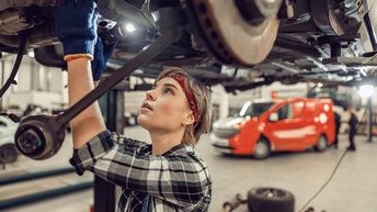 Eine Person mit Handschuhen steht unter einem über ihr fixierten Auto. Die Person hält in der rechten Hand eine Taschenlampe und blickt auf den Unterboden des Autos. Im Hintergrund stehen weitere Autos im Raum