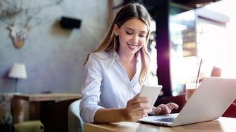 Person mit langen braun-blonden Haaren sitzt lächelnd bei einem Tisch in einem Restaurant und blickt dabei auf ein Smartphone, während auch mit einem Laptop gearbeitet wird, daneben steht ein Getränk am Tisch