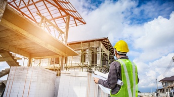 Rückenansicht einer Person mit Helm, Warnweste und Architektenplan, die zu einer Baustelle eines Gebäudes blickt. Daneben ist eine weitere Baustelle eines Gebäudes