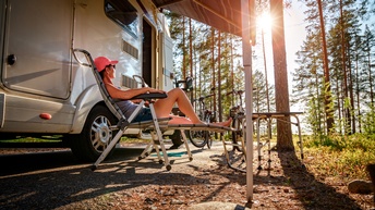 Eine Person mit Kappe, kurzer Hose und Oberteil sitzt in einem Campingstuhl vor einem Wohnwagen. Der Wohnwagen steht in einem Wald. Der Person blickt zur am Himmel stehenden Sonne. Neben der Person stehen Fahrräder